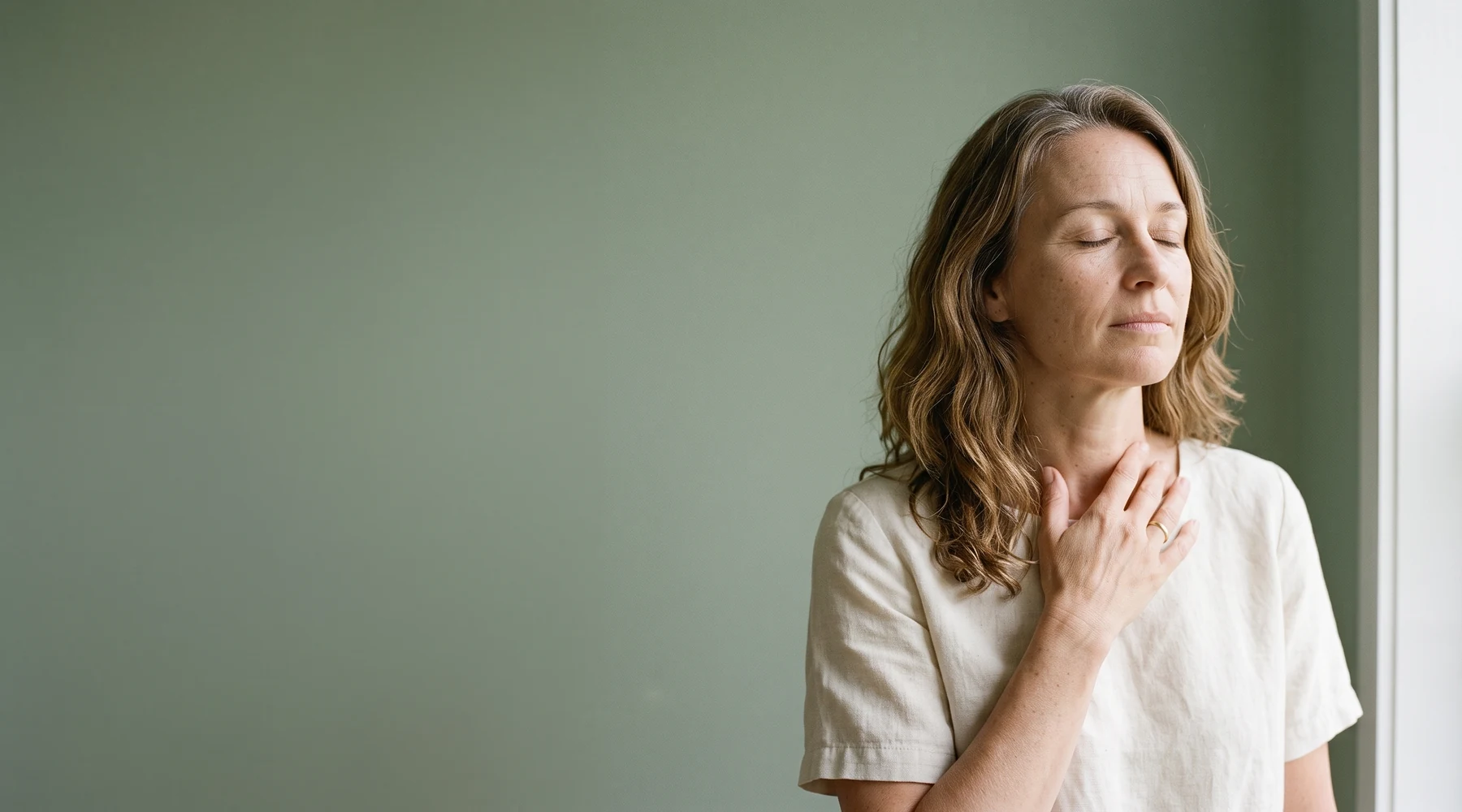 Femme de 48 ans les yeux fermés, la main sur la clavicule, devant un mur sauge